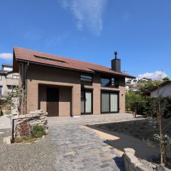 赤瓦の屋根から薪ストーブの煙突が見えている。玄関に続くアプローチに石が敷かれ、石を積んだ門塀に設置された表札を門灯が照らしている
